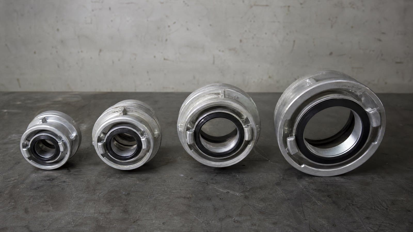 Four aluminium Storz fire hose couplings of increasing size in a row on a grey workshop bench