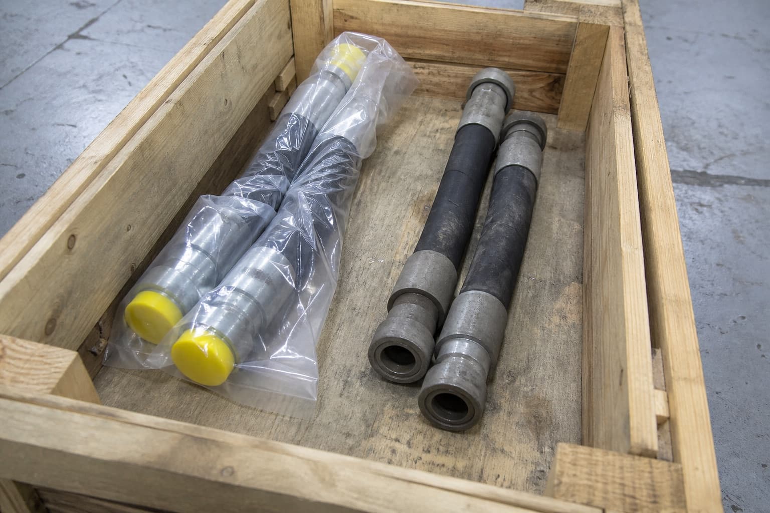 An opened timber crate holding two components sealed in polythene with bright caps fitted on their ends, beside two bare ones with open uncapped ends dulled by dust
