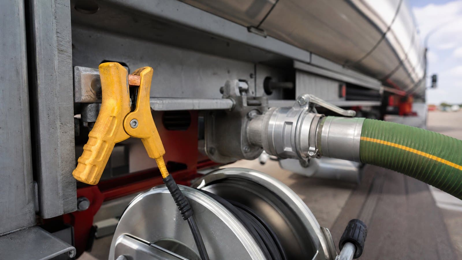 Yellow earthing clamp on a fuel tanker bracket beside the aluminium coupling of a green fuel hose and a grounding reel