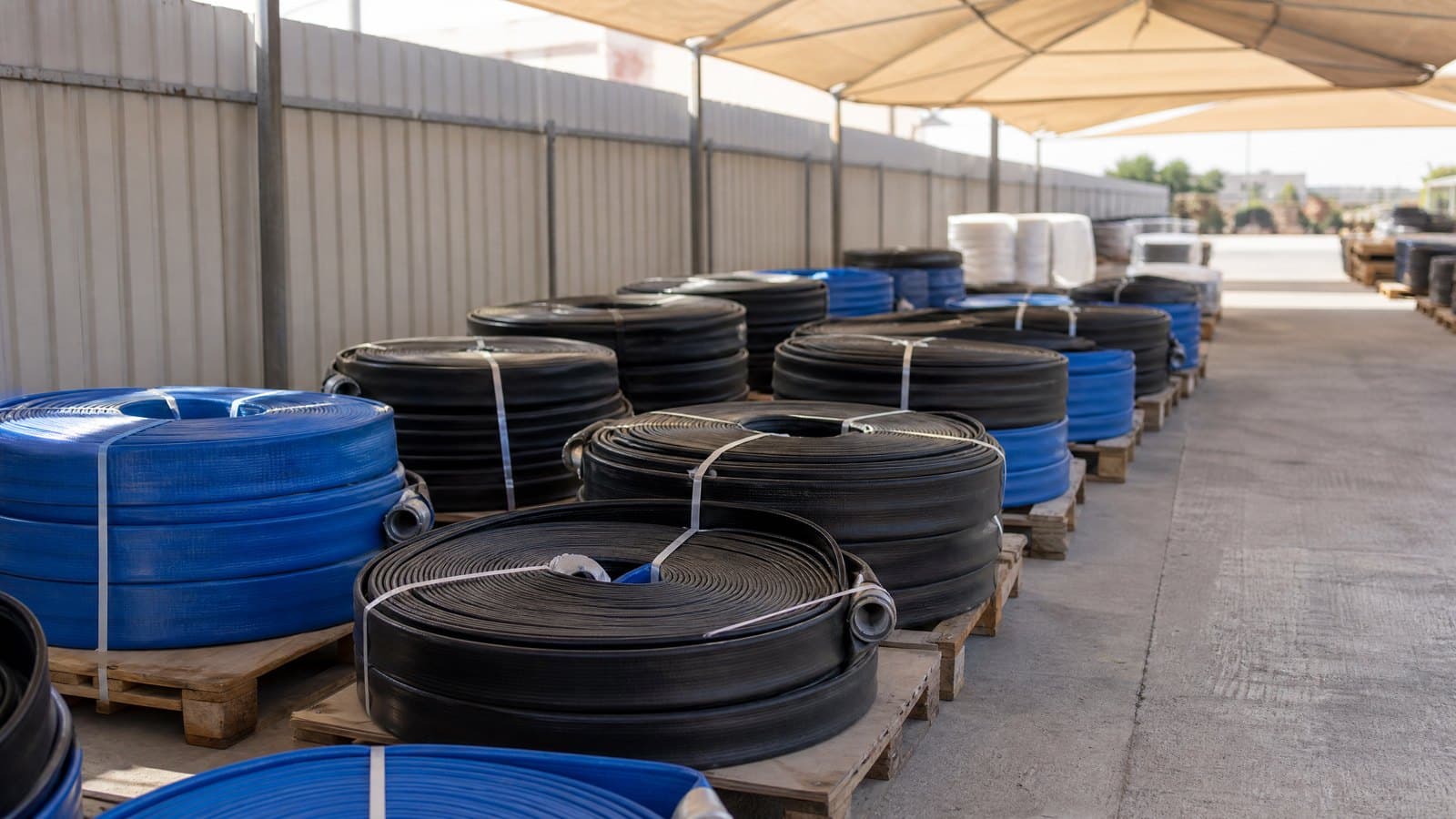 Coils of black and blue lay flat hose strapped on pallets under a shade canopy in a contractor yard