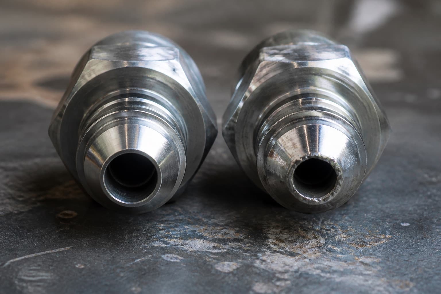 Two hydraulic fittings side by side on a workbench, the cone on the left cleanly machined and the one on the right crushed and burnished at its tip