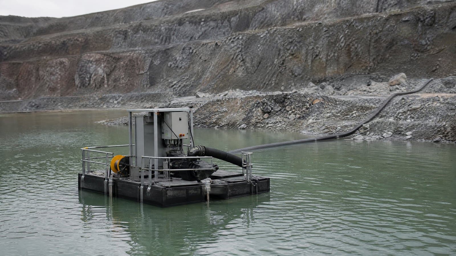 Pump on a floating pontoon in a mine pit-bottom pond with a black discharge hose running to the rock face
