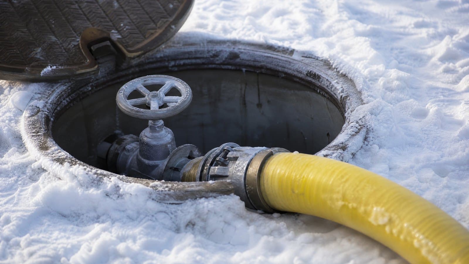 Open snowmaking hydrant pit on a ski slope with a valve and a yellow hose coupled to it, snow around