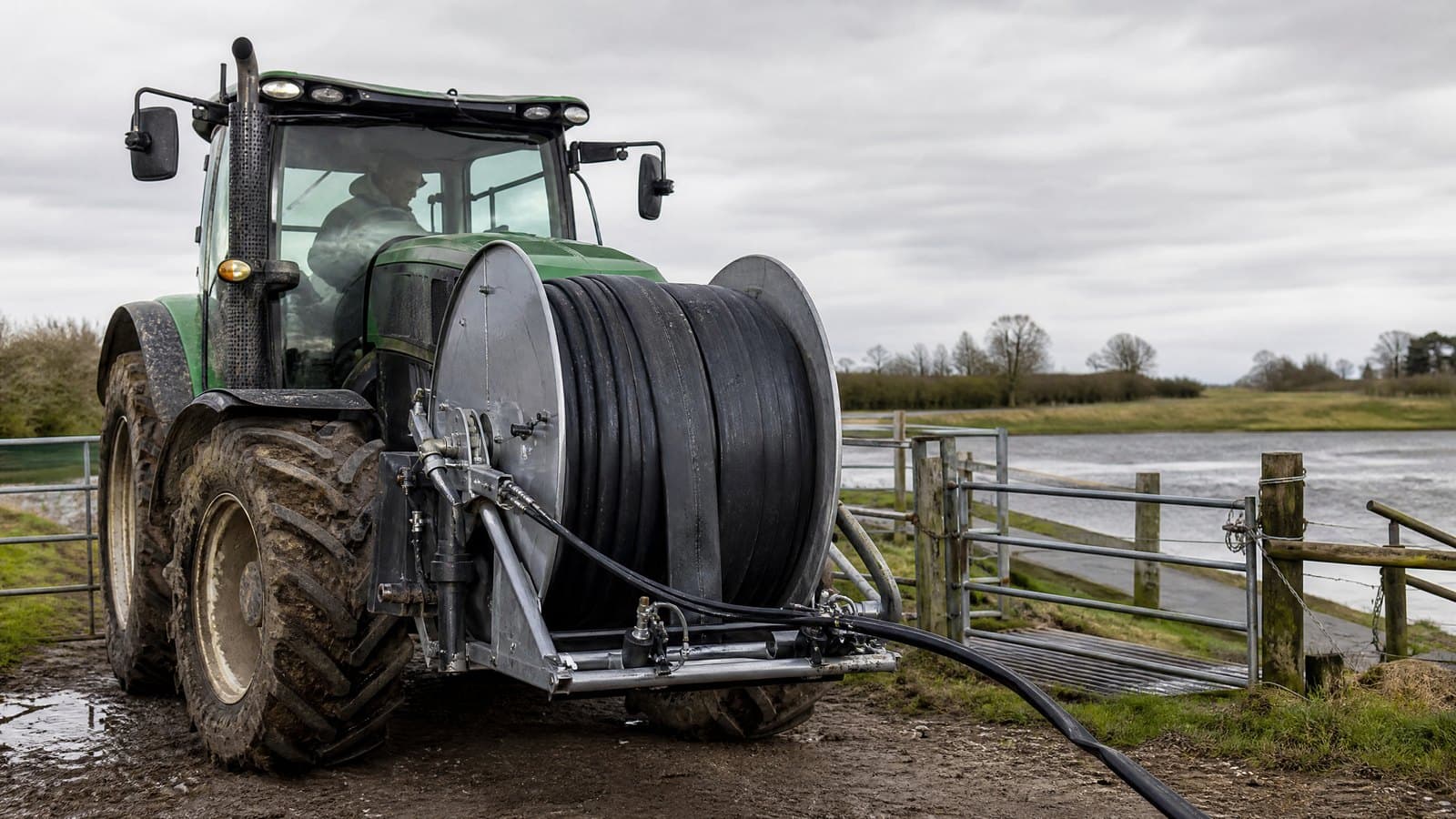 Tractor with a front-mounted hydraulic reel of black lay flat supply hose at a field gate beside a lagoon