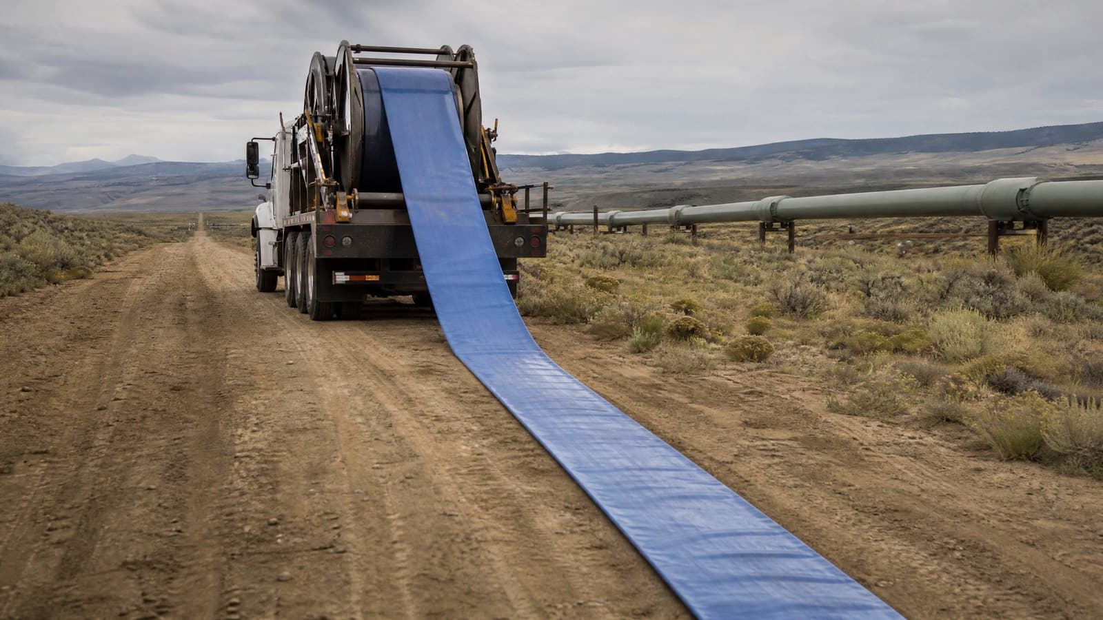 Truck laying blue lay flat hose from a drum along a dirt road beside a pipeline