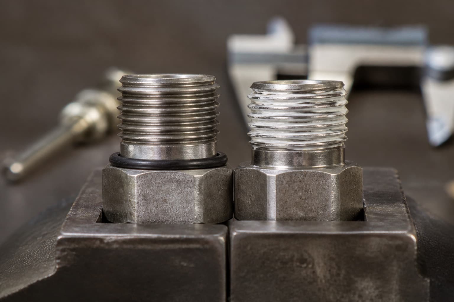 A parallel-thread fitting with a bonded seal beside a tapered-thread fitting wound with sealing tape, stood side by side in a bench vice