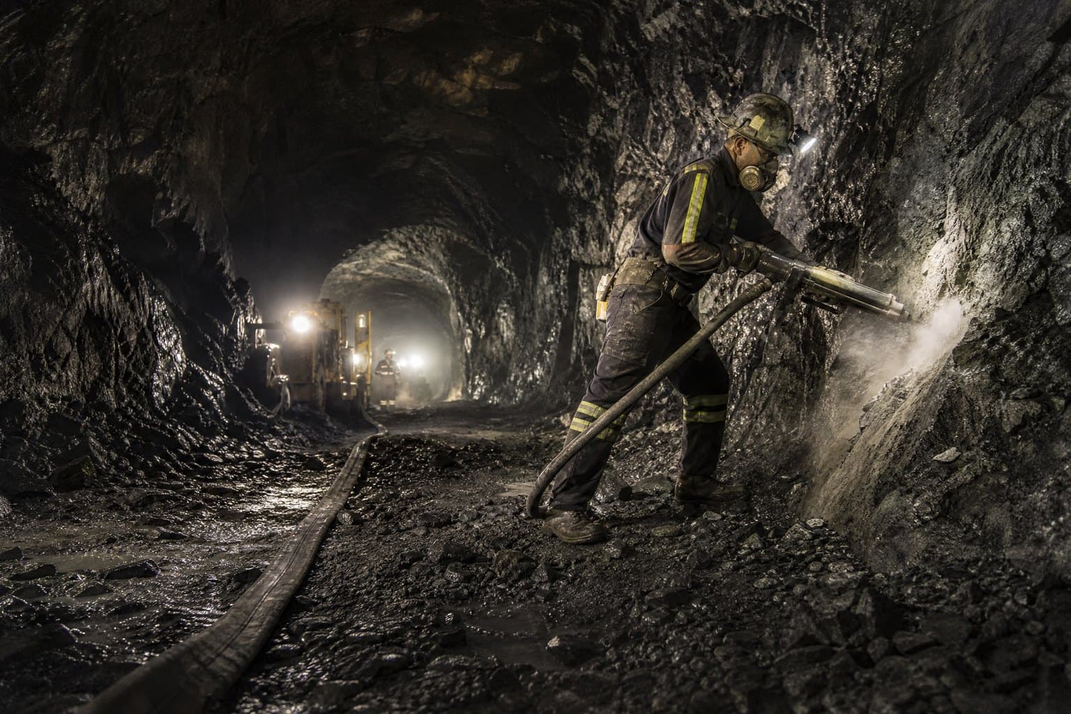 Miner operating a pneumatic rock drill in an underground tunnel, fed by a hose running along the tunnel floor