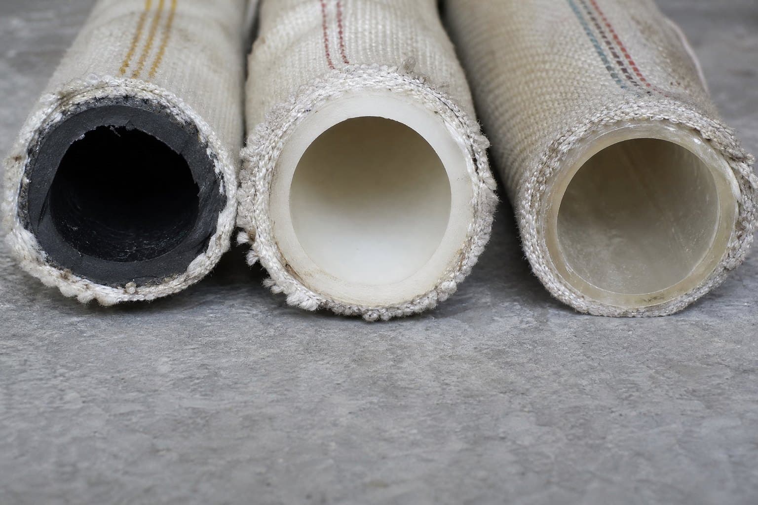 Cut ends of three woven-jacket fire hoses showing black rubber, off-white rubber and thin translucent polyurethane liners