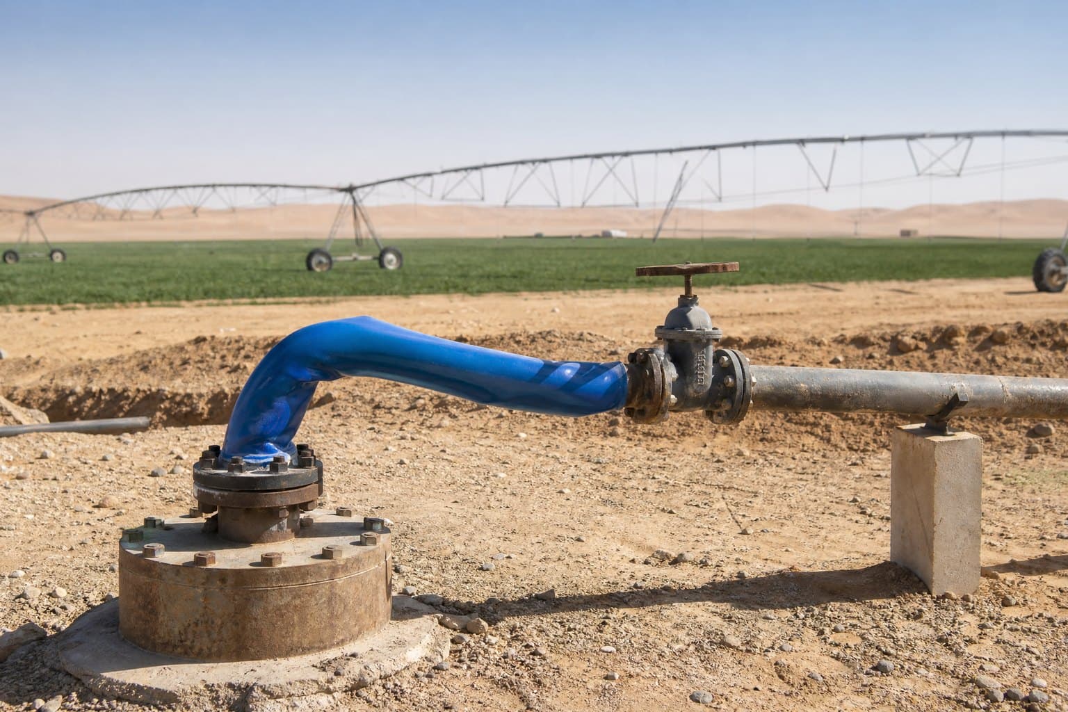 Blue flexible riser hose rising from a borehole well head to a valve, centre-pivot irrigated fields behind