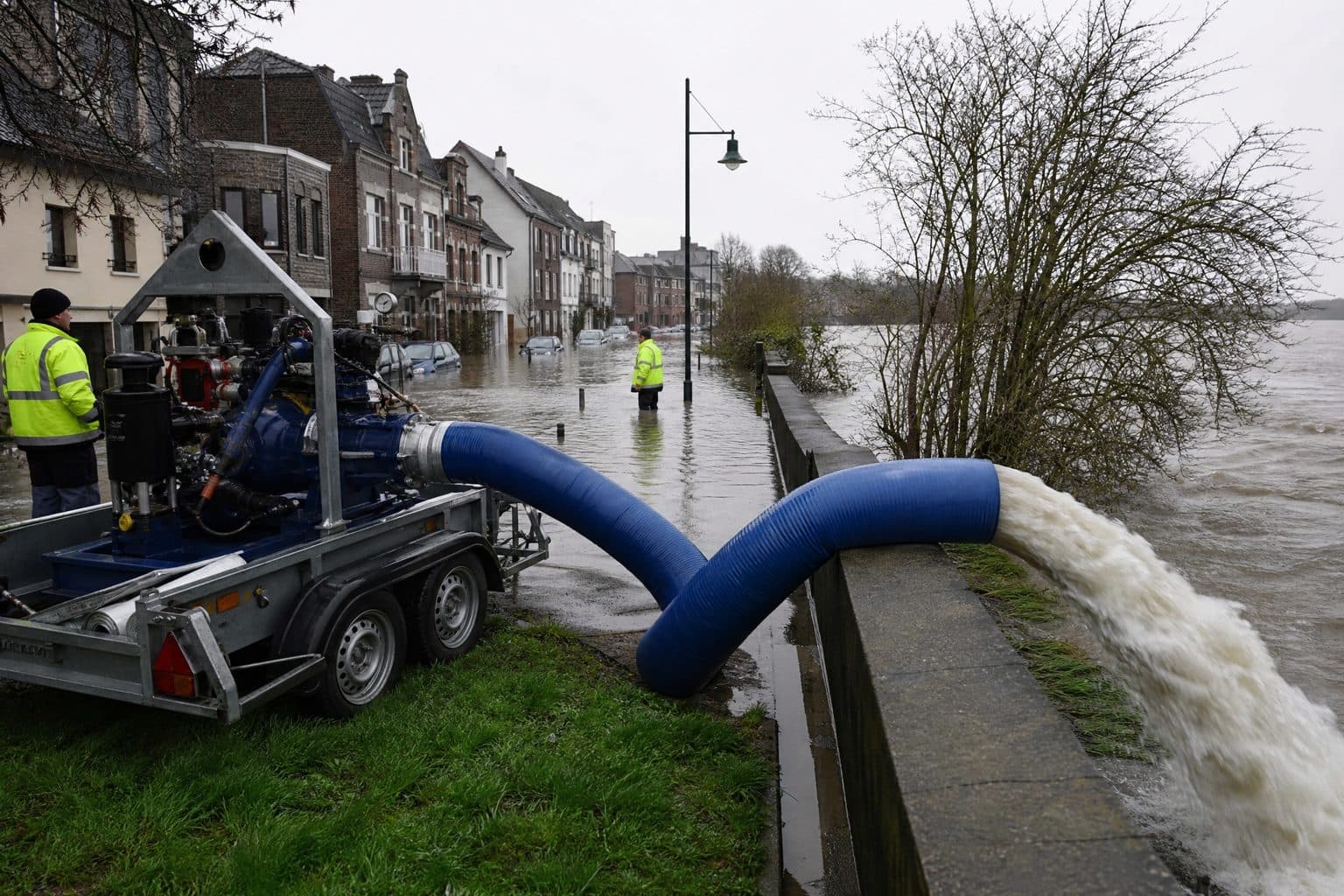 Trailer pump on a flooded town street discharging through a large blue lay flat hose over a flood wall into a river
