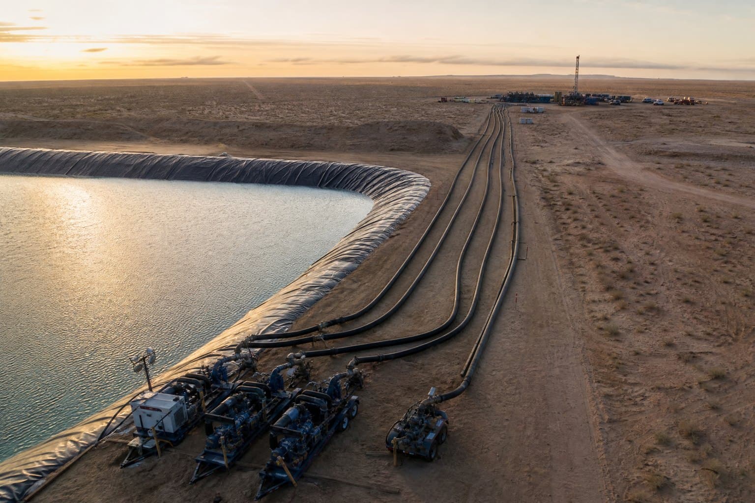 Aerial view of large black lay flat hoses running from transfer pumps at a lined desert pond towards a distant well pad