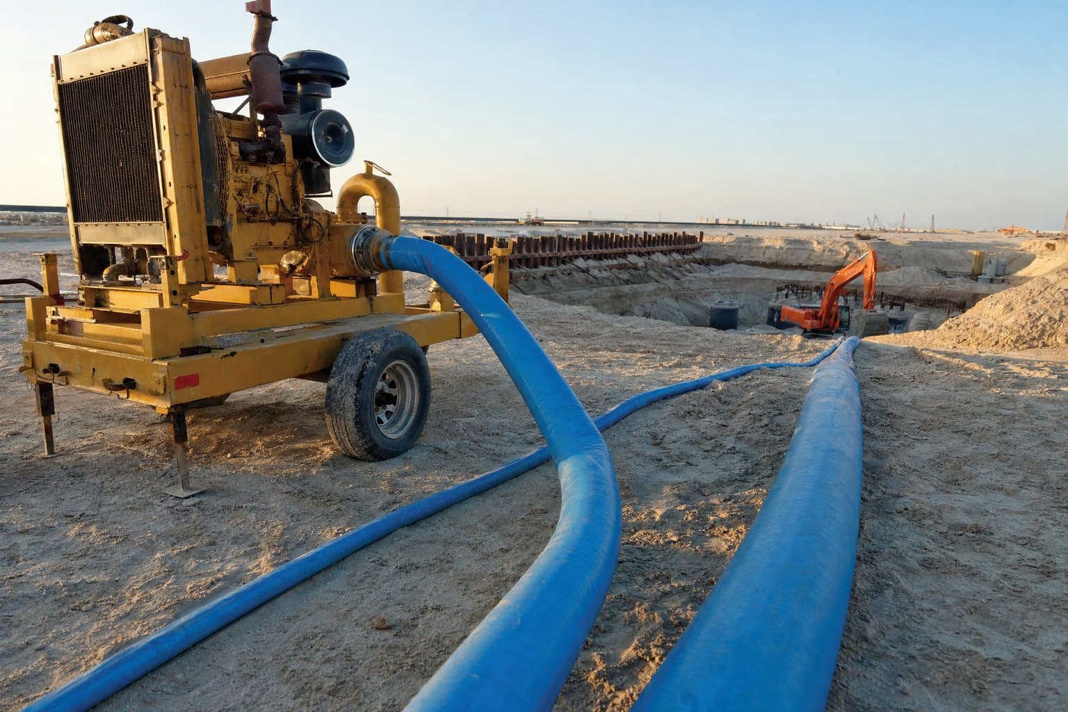 Blue lay flat hose lines running from a trailer-mounted diesel pump across a sandy construction site to an excavation