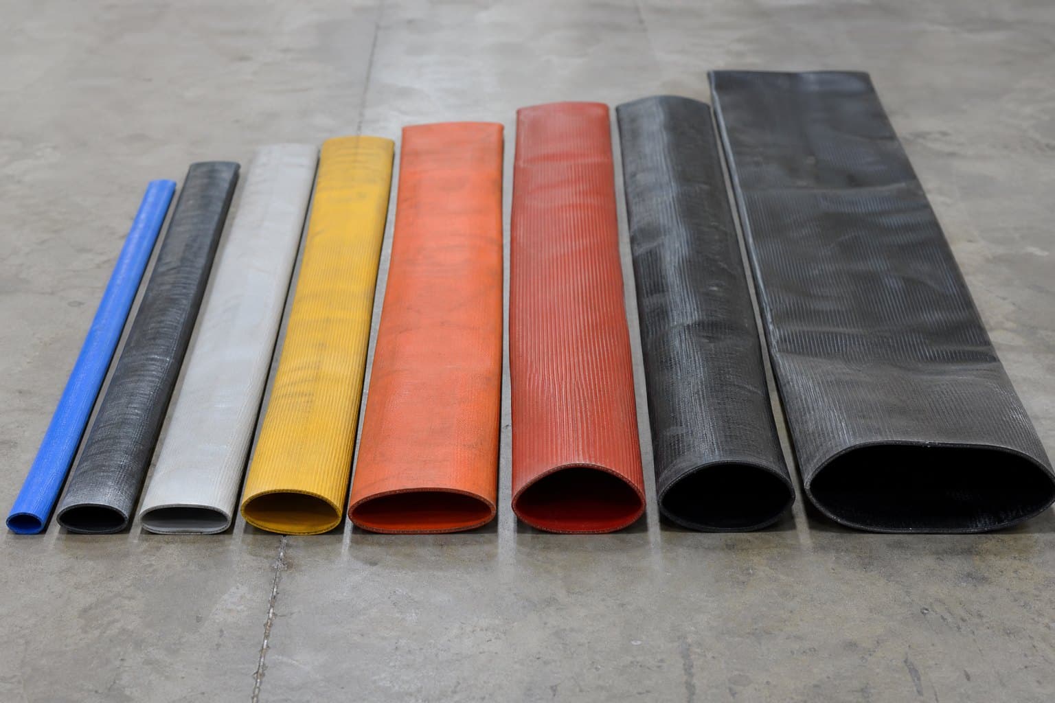 Flattened lay flat hose ends of increasing width in blue, grey, yellow, orange, red and black laid side by side on concrete