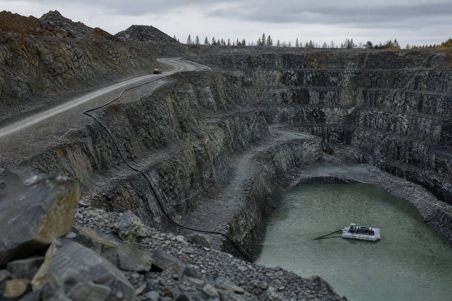 Deep open-pit mine with terraced rock benches and a pump pontoon in the pit-bottom pond, hose running up the benches