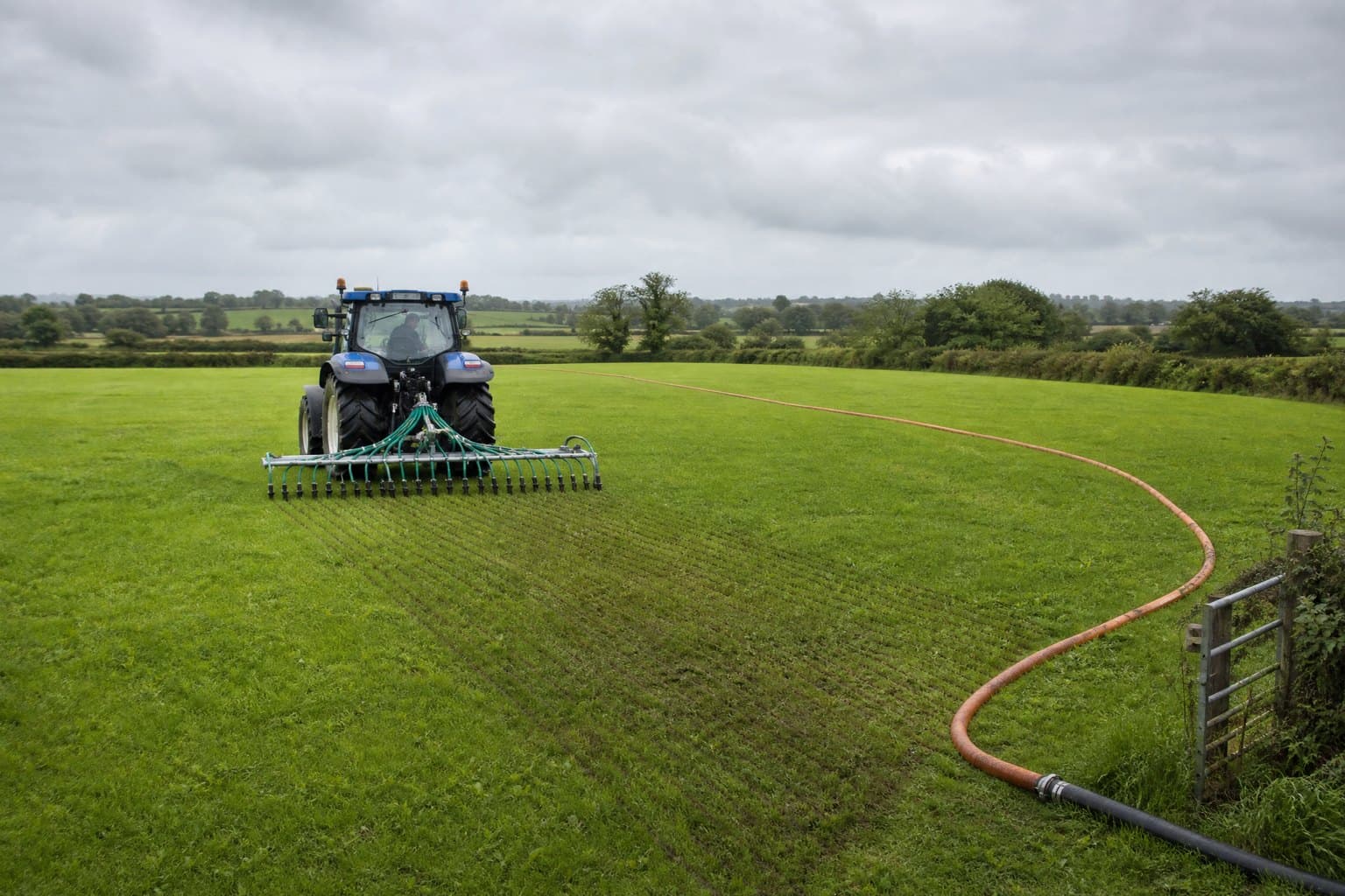 Tractor with a trailing-shoe boom on grassland pulling an orange drag hose that joins a black supply hose at the field gate