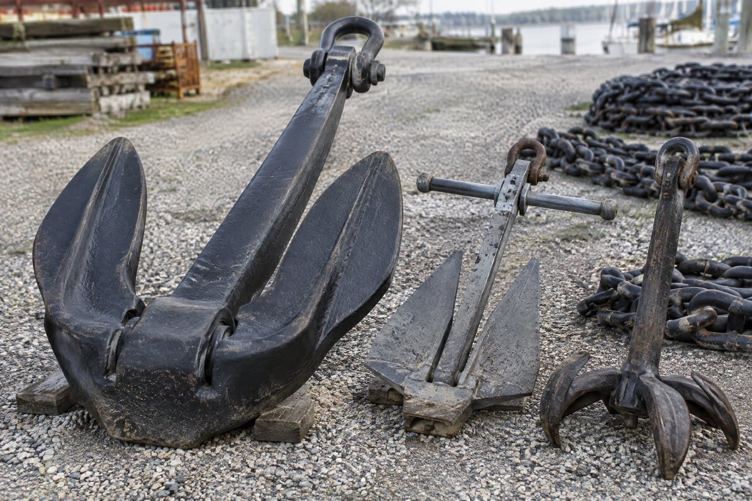 A black stockless anchor with hinged flukes, a Danforth anchor and a claw anchor on gravel in a marine yard, anchor chain behind