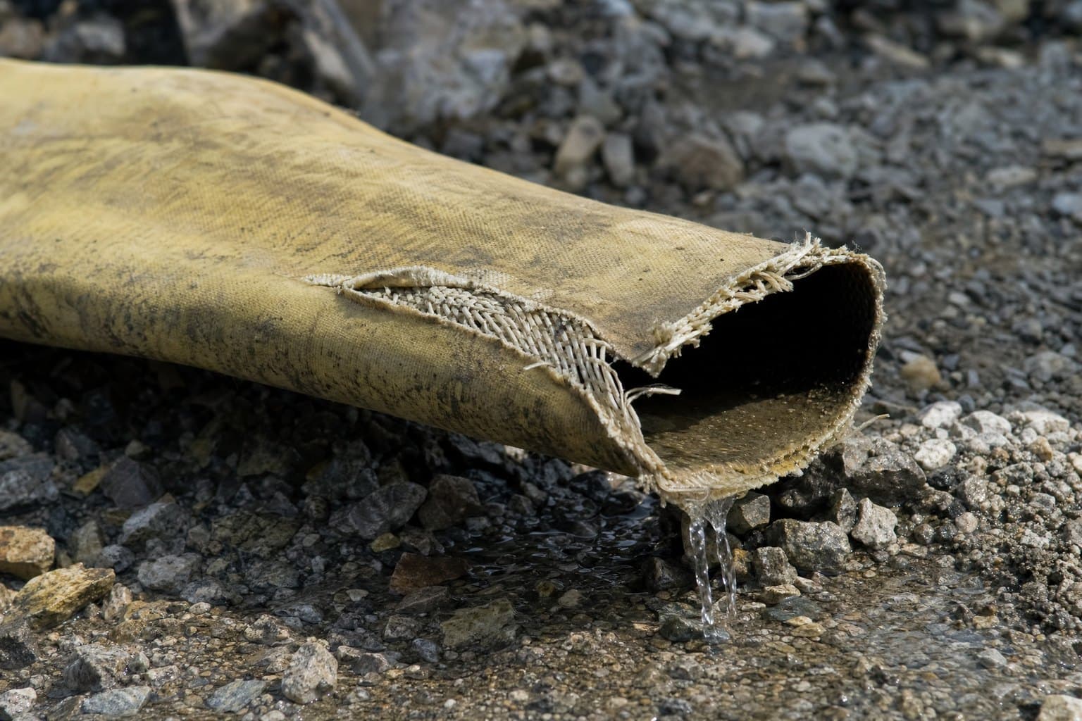 Worn lay flat hose on gravel with a split along the fold and exposed woven jacket fibres